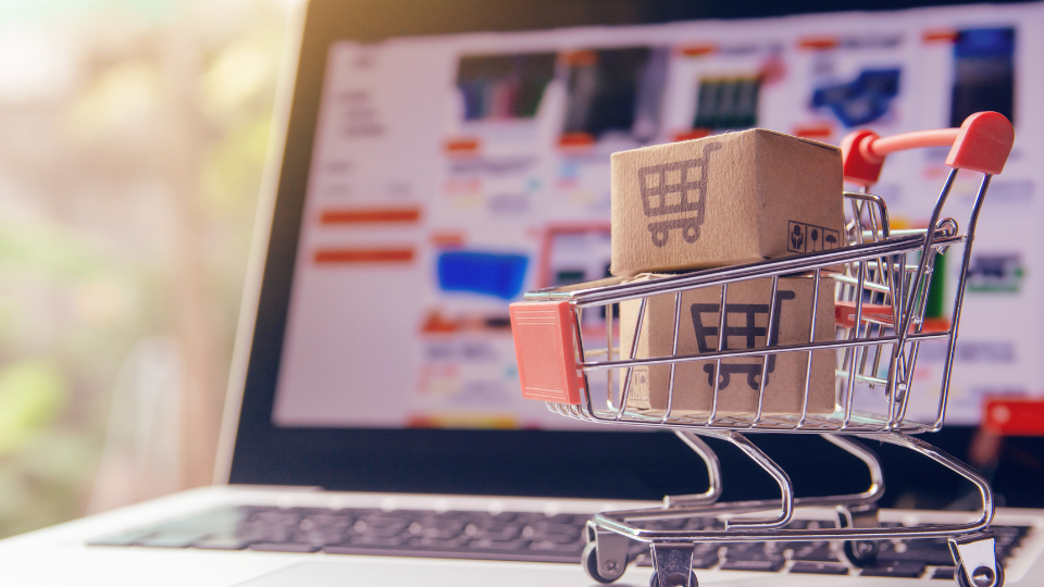 A mini shopping cart filled with boxes sits on top of a laptop which is out of focus in the background